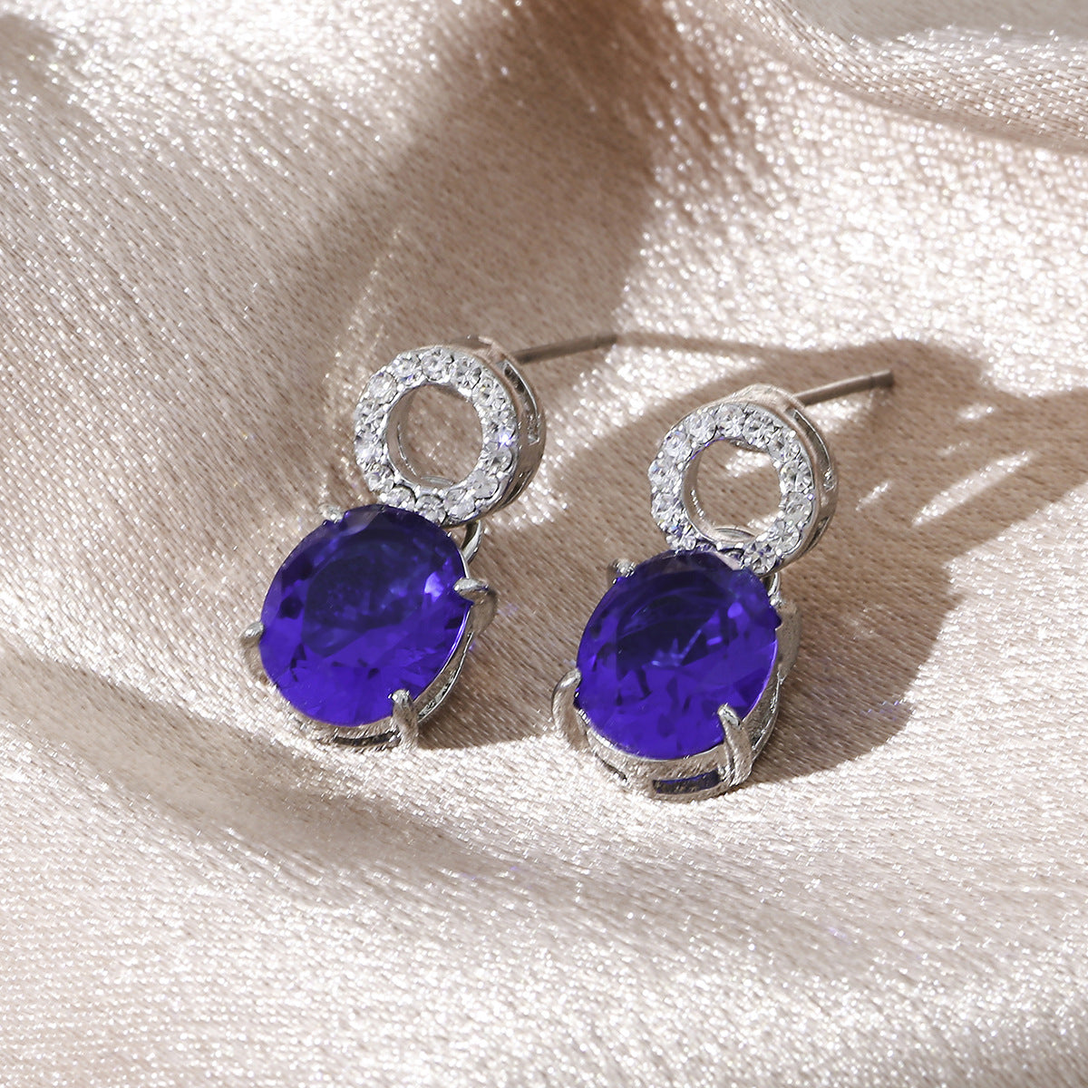 luxury royal blue oval cut sapphire drop earrings with shimmering micro paved crystal studs on a silk background