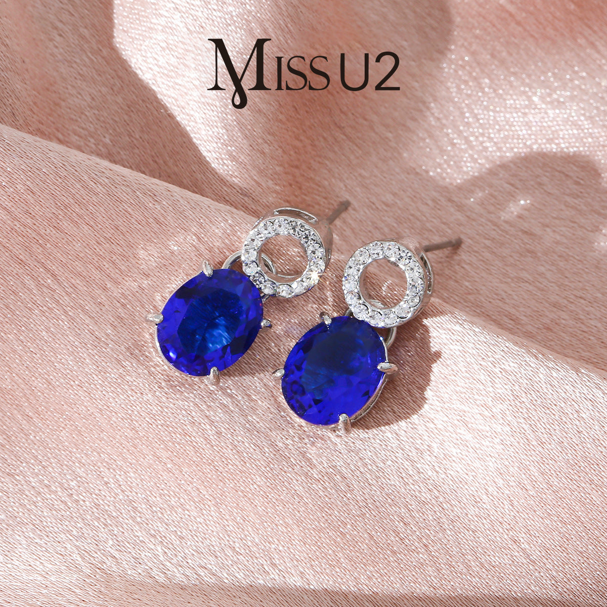 luxury royal blue oval cut sapphire drop earrings with shimmering micro paved crystal studs on a silk background